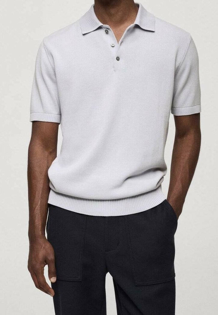 Homme portant un polo en maille gris clair à manches courtes avec trois boutons et un pantalon noir, debout avec une main dans la poche.