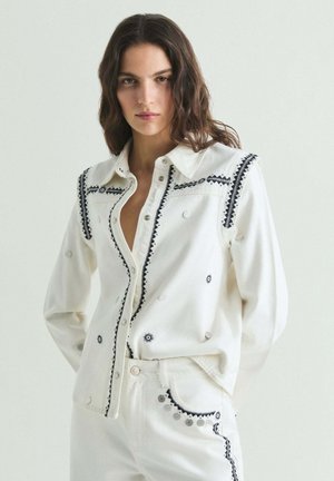 Mujer de pie con camisa blanca abotonada y pantalones de pierna ancha con detalles bordados en negro, combinados con sandalias negras abiertas en la punta.