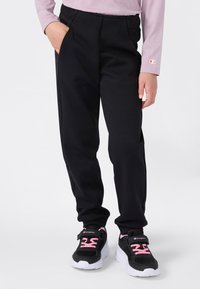 Kind trägt schwarze Hosen, schwarze und pinke Sneakers und ein langärmliges lila T-Shirt mit der Hand in der Tasche vor einem weißen Hintergrund.