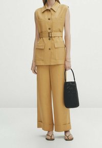 Senapsgul ärmlös skjorta med krage, knäppning fram och bälte. Vida byxor och en svart vävd handväska kompletterar outfiten.