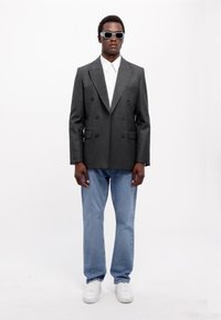 Grå dobbeltknappet blazer over en hvid skjorte med krave, parret med lyseblå jeans og hvide sneakers. Accessoriseret med grå solbriller.