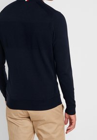 Granatowy sweter z dzianiny z teksturowanymi poziomymi paskami, ze ściągaczami przy mankietach i u dołu, z małym czerwono-białym akcentem na tylnym dekolcie.