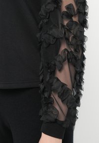 Suéter negro con un cuerpo sólido y mangas de tela transparente adornadas con acentos de tul negro con volantes. Los puños ajustados completan el diseño.