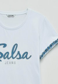 Camiseta blanca de manga corta con texto azul decorado "Salsa" y pequeña inscripción "Jeans", con ribete de cadena y tela a lo largo del borde de la manga.