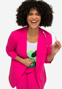 Blazer ajusté rose vif et pantalon assorti, avec une chemise blanche ornée d'un design graphique coloré. Le mannequin porte des lunettes et sourit.