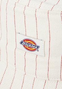 Biely klobúk s krátkym okrajom s červenými vertikálnymi pruhmi, ktorý má na boku obdĺžnikový vyšitý logotyp Dickies. Hladká textúra látky.