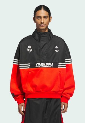 Joven con cabello largo vestido con una chaqueta negra y roja que presenta franjas blancas, un gráfico de rosa, logotipos de Adidas y el texto "CHAVARRIA" en el pecho.