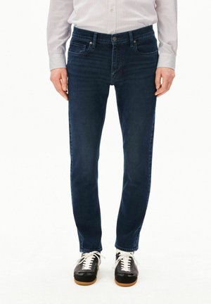 Persoon staat rechtop, draagt donkerblauwe jeans, zwarte sneakers met witte veters en een licht lang shirt met lange mouwen. Handen ontspannen naast het lichaam.