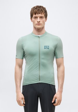 Άνδρας που φοράει εφαρμοστή ανοιχτοπράσινη κοντομάνικη ποδηλατική φανέλα με φερμουάρ μπροστά και μαύρο σορτς, στέκεται μπροστά σε απλό φόντο.