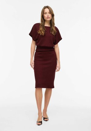 Žena stojaca, oblečená v bordovom tričku s krátkym rukávom a zodpovedajúcej sukni po kolená, s čiernymi sandálmi s otvorenou špičkou, na bielom pozadí.