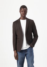 Brauner Blazer mit zwei vorderen Taschen, Ein-Knopf-Verschluss und Reverskragen, getragen über einem weißen T-Shirt und kombiniert mit blauer Jeans.