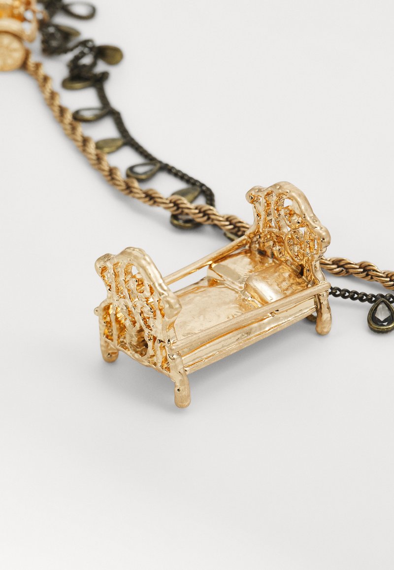 Colgante miniatura de banco en tono dorado con detalles intrincados, que presenta un respaldo ornamentado y un asiento liso, unido a una cadena con acentos decorativos.