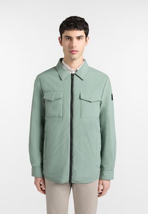 Ung mand, der vender fremad iført en lysegrøn lynlåstrøje med klaplommer og beige bukser mod en ensfarvet hvid baggrund.