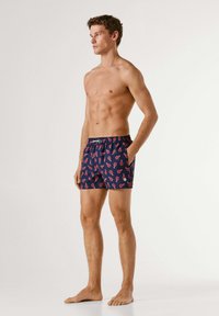 Skjorteløs mand stående barfodet i marineblå badeshorts med rød hummerprint, hænderne i lommerne, kiggende mod venstre mod en ensfarvet baggrund.