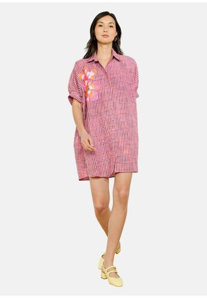 Frau geht in einem pink karierten Hemdkleid mit orangem Blumenmuster auf der Schulter, trägt gelbe Mary Jane Pumps.