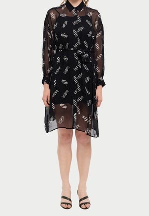Femme portant une robe noire transparente avec des motifs géométriques blancs, manches longues, col, ceinture à la taille et talons ouverts noirs.