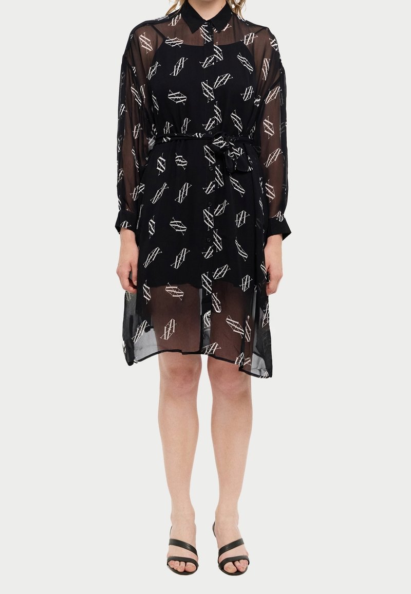 Femme portant une robe noire transparente avec des motifs géométriques blancs, manches longues, col, ceinture à la taille et talons ouverts noirs.