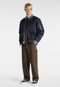 Marinebomberjacke mit gerippten Bündchen, hellblauen Akzenten; über einem braun-lila gestreiften Pullover getragen, kombiniert mit braunen Hosen und Sneakers.