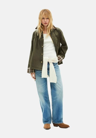 Chaqueta verde oliva con puños de cuadros, camiseta blanca y vaqueros azul claro. Tela color marfil atada a la cintura. Botas marrones completan el conjunto.