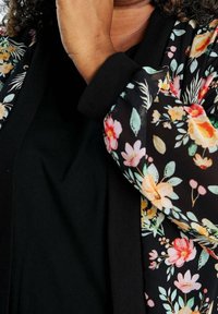 Persoon met een zwart jasje met bloemenpatroon houdt zijn kin vast, met krullend haar en een zwart binnenstuk zichtbaar.