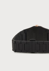 Montre noire avec une finition mate, dotée d'un bracelet à maillons rectangulaires, d'un boîtier rond et d'une couronne en or rose sur le côté.