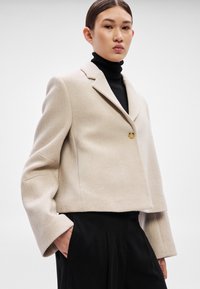 Μπεζ cropped σακάκι με φαρδιά μανίκια, κλείσιμο με ένα κουμπί και λεπτή υφή. Φορεμένο πάνω από μαύρο ζιβάγκο, σε συνδυασμό με μαύρο παντελόνι.