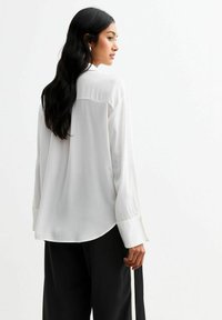 Witte blouse met lange mouwen, gemaakt van een gladde stof en met een ontspannen pasvorm. Heeft een achterpand en een gebogen zoom, gecombineerd met zwarte broeken.