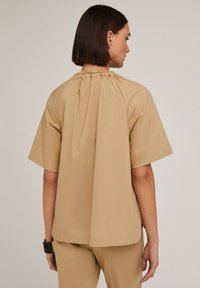 Top beige in cotone a maniche corte con scollatura arricciata, vestibilità rilassata e superficie liscia, visto da dietro, abbinato a pantaloni coordinati.