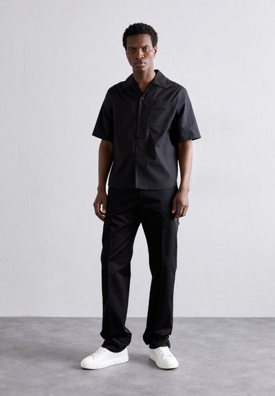 Chemise noire à manches courtes avec fermeture à boutons et poche, associée à un pantalon noir. Le modèle porte des baskets blanches. Design simple, sans motifs.
