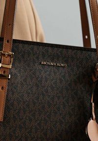 Sac à main brun à motifs avec quincaillerie dorée, doté de deux anses réglables et d'un logo subtil. Le matériau texturé ajoute de la profondeur.