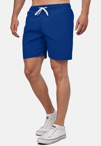 Blauwe zwembroek met een elastische tailleband en een witte trekkoord. Glad stof, knielengte ontwerp, met een klein logo detail.