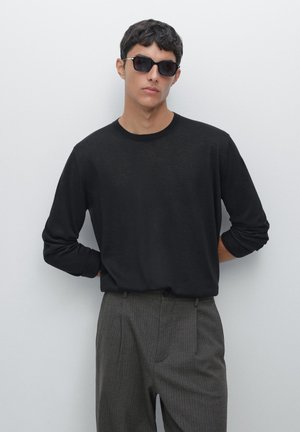 Schwarzes Langarmshirt aus weichem Stoff mit Rundhalsausschnitt, kombiniert mit grauen gestreiften Hosen. Die Accessoires umfassen dunkle Sonnenbrillen.