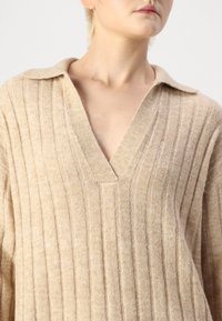 Beige bordázott pulóver V-kivágással és széles gallérral, puha, texturált anyagból készült. Függőleges csíkokkal díszítve.