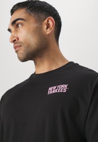 Camiseta de algodón negra con un logo morado de "NEW YORK YANKEES" en el lado izquierdo del pecho. Diseño clásico de cuello redondo con mangas cortas.