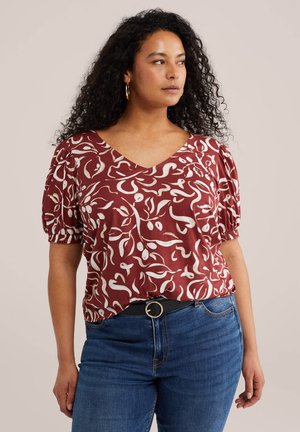 Žena s kovrčavom kosom nosi naušnice u obliku ringova, bordo bluzu kratkih rukava s uzorkom, plave traperice i crni remen s okruglom kopčom.