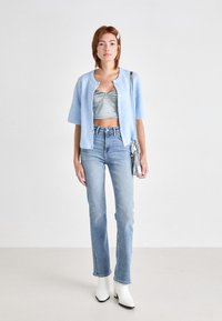 Cardigan azzurro chiaro a coste con maniche corte, indossato sopra un top grigio corto, abbinato a jeans azzurri chiari e stivali bianchi alla caviglia.
