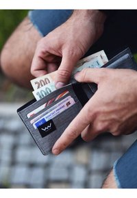 Gray fabric wallet with black tag, holding cash and cards with visible denominations of 100 and 200 euros. Hands partially visible.