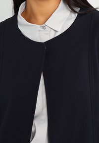 Laivastonsininen neuletakki, jossa pyöreä pääntie, valmistettu sileästä kankaasta. Siinä on piilotettu etukiinnitys, ja se on kerroksittain valkoisen kauluksellisen paidan päällä.