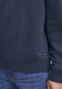 Navyblauwe sweatshirt van zacht materiaal, met ribgebreide manchetten en onderkant. Kleine label onderaan met tekst. Draagt met blauwe spijkerbroek.
