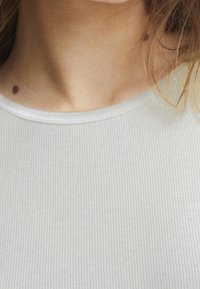 Světle šedý žebrovaný top bez rukávů s výstřihem, který má jemnou texturu, úzký střih a zaoblený límec s decentním prošíváním.