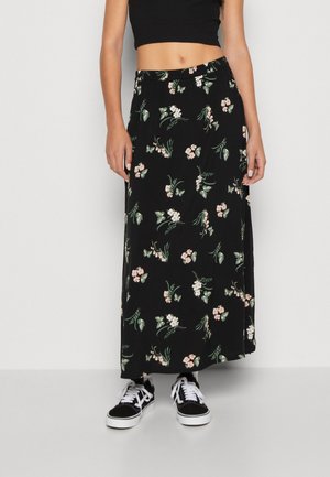 Kvinde iført en sort maxi nederdel med blomsterprint, sort crop top og sorte og hvide sneakers, stående mod en ensfarvet baggrund.