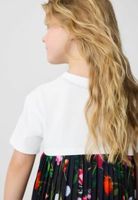 Valge lühikeste varrukatega topp, millele on kombineeritud pleeritud must lilleline seelik, kus on erksad värvid. Pikk laineline juuksed langevad õlale.