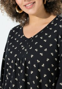Zwarte v-hals tuniek met wit bloemenpatroon, lange mouwen en een geplooid ontwerp, gemaakt van zachte stof. Grote ronde oorbellen zichtbaar.