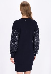 Sötét tengerészkék fonott pulóverruha érdes, flitteres ballonujjakkal, bordás mandzsettával és szegéllyel. Simább test, finom hátsó logó részlettel.