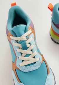 Helles blaues Sportschuh aus Wildleder und Mesh-Material, mit braunen Lederakzenten, weißen Schnürsenkeln und einer mehrfarbigen Sohle.