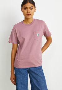 T-shirt di cotone rosa con colletto rotondo, maniche corte e taschino sul petto con un logo. Abbinato a pantaloni in denim a gamba larga blu.