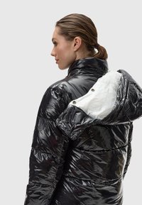 Zwarte pufferjack gemaakt van glanzend synthetisch materiaal met een witte binnenvoering, drukknoopsluitingen en een capuchon met een opvallend ontwerp.