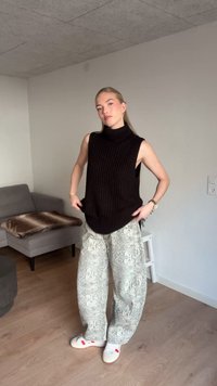 Svart stickad ärmlös polotröja med ribbad textur, kombinerad med ljusgrå, ormskinnsmönstrade vida byxor och vita sneakers.