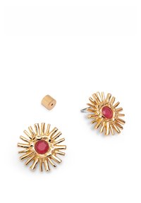 Pendientes en tono dorado con un diseño de explosión solar, que presentan una gema roja central. Incluyen una pieza cilíndrica pequeña adicional.