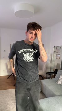 Čierne grafické tričko s vyblednutou textúrou a bielym dizajnom cheruba. Šedé voľné džínsy, viditeľná tetovaná ruka, stojí v svetlej miestnosti.